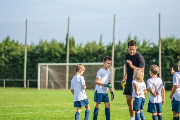 Group of children in sports kits gathered on a grass football pitch with an adult, near a goal in the background.
