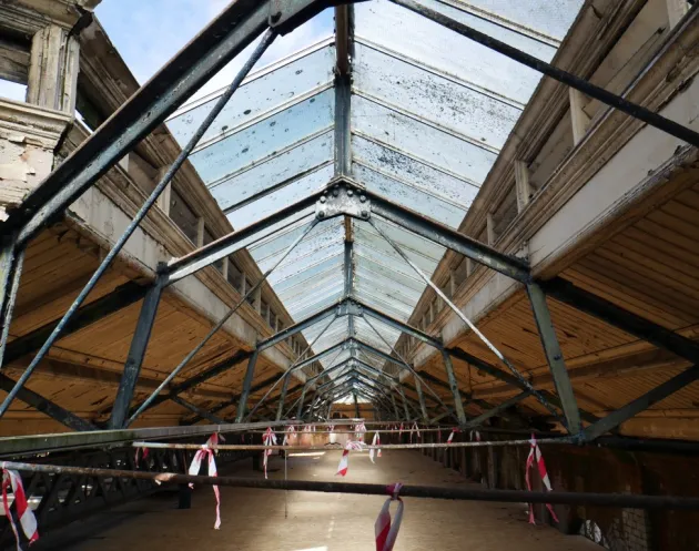 Interior view of the Iveagh Markets showing the long span metal roof structure with semi‑glazed panels, cast iron trusses and temporary safety barriers below.
