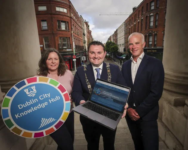 Three people stand together. The woman on the left holds a 'Dublin City Knowledge Hub' sign and the man in the middle holds a laptop. 