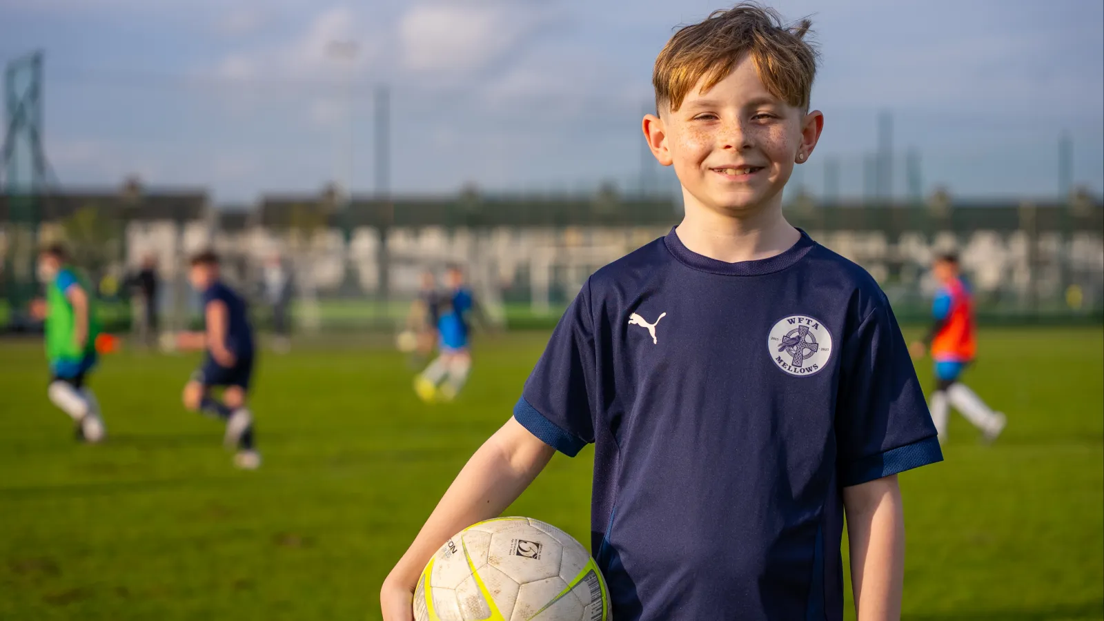 young boy holds football as match is played behind him